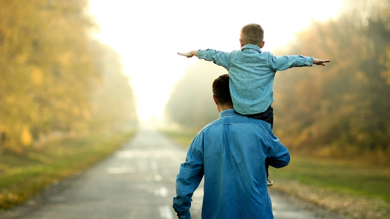 Ein Vater trägt sein Sohn auf den Schultern und sie stehen beide auf einem Kiesweg